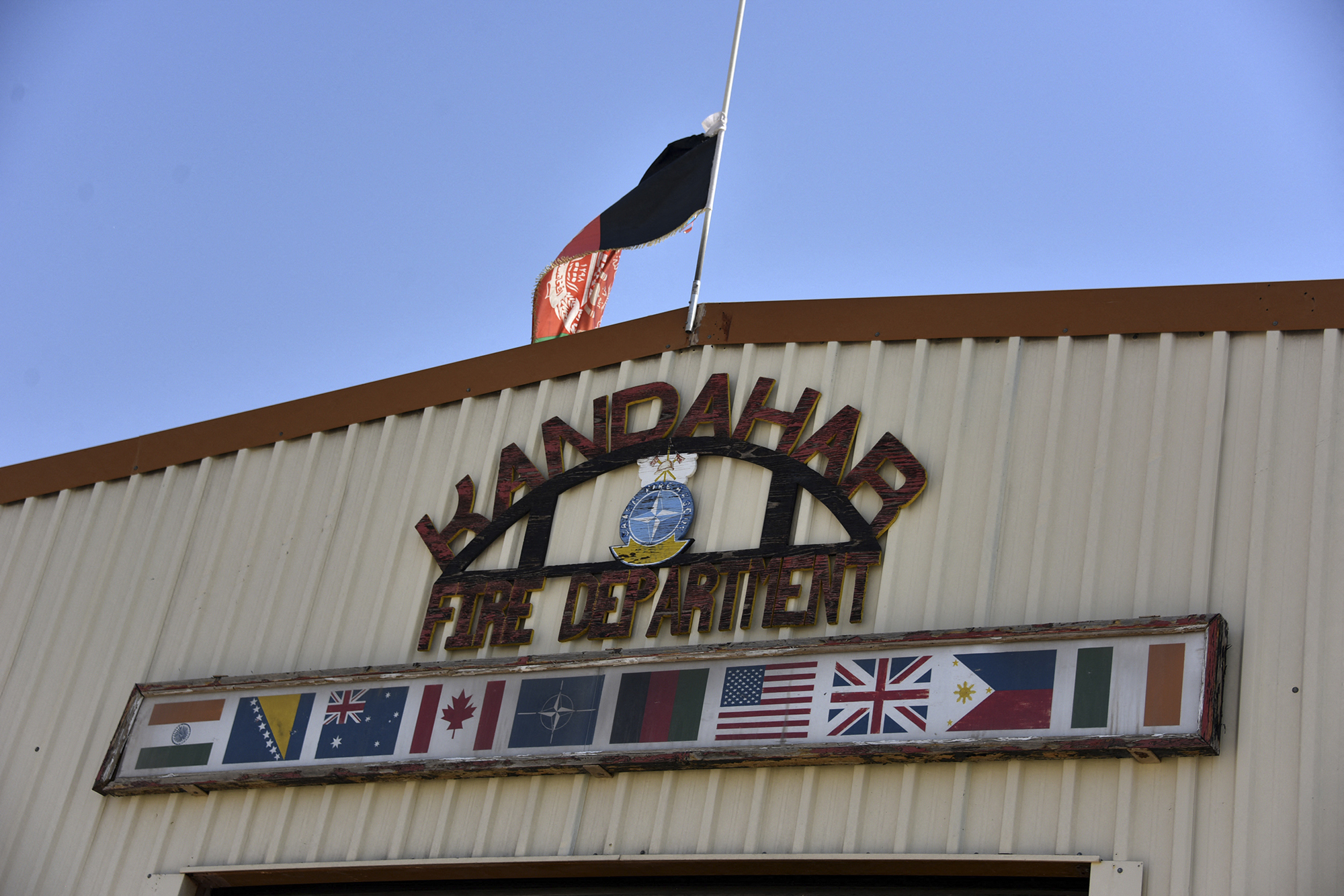 The Afghanistan flag flies over a sign that reads "Kandahar Fire Department" on the side of a building. The flags of NATO nations are displayed under the sign.